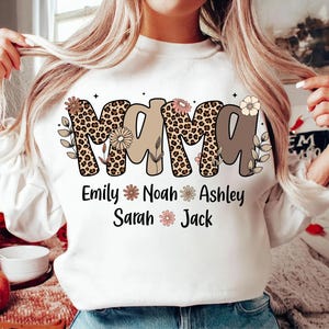 May include: White sweatshirt with the word "MAMA" in a mix of leopard print and brown lettering, decorated with floral accents. Below the word "MAMA" are the names Emily, Noah, Ashley, Sarah, and Jack, each with a flower symbol.
