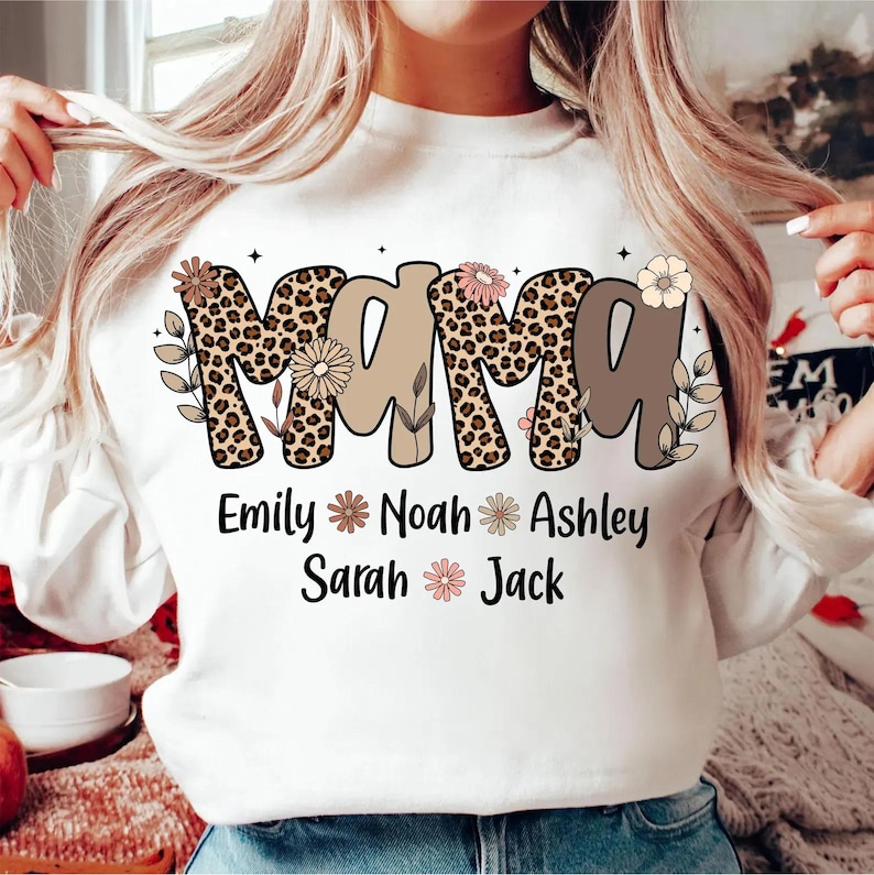 May include: White sweatshirt with the word "MAMA" in a mix of leopard print and brown lettering, decorated with floral accents. Below the word "MAMA" are the names Emily, Noah, Ashley, Sarah, and Jack, each with a flower symbol.