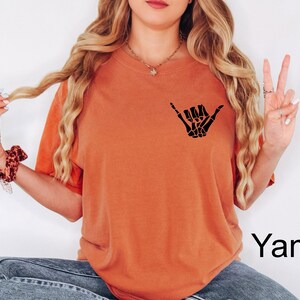 Pode incluir: Uma camiseta laranja queimada com uma m&atilde;o de esqueleto preta fazendo um sinal shaka. O texto "Yam" &eacute; vis&iacute;vel no canto inferior direito da imagem.