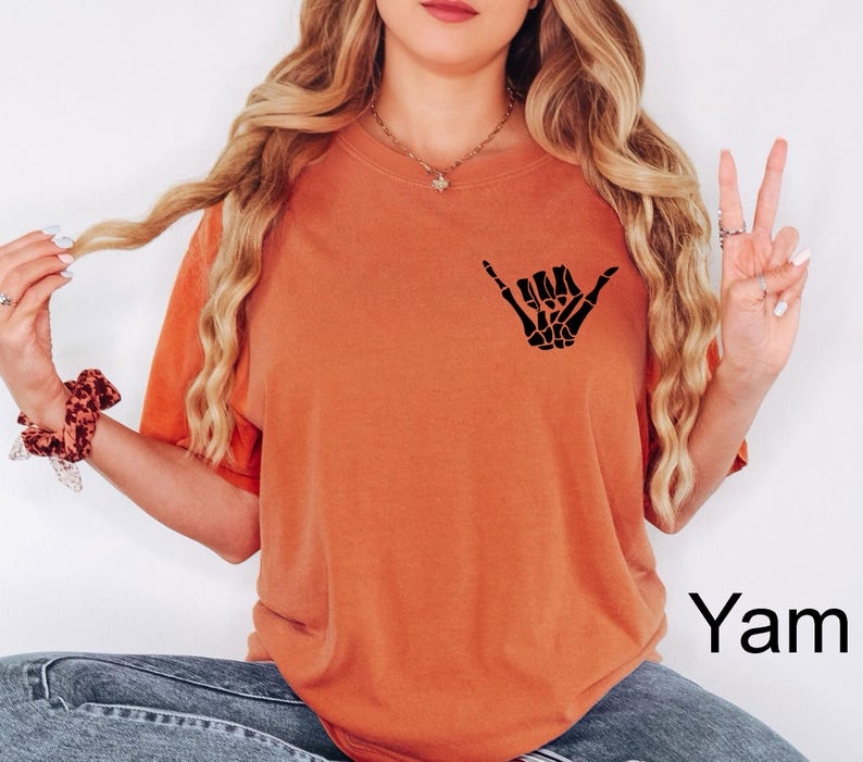 Pode incluir: Uma camiseta laranja queimada com uma m&atilde;o de esqueleto preta fazendo um sinal shaka. O texto "Yam" &eacute; vis&iacute;vel no canto inferior direito da imagem.