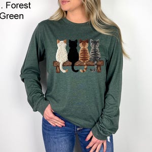 May include: Forest green long-sleeved top with an embroidered design of four cats on a wooden bench. The cats are white, black, brown, and grey. The shirt has a crew neck and is made of a soft material.