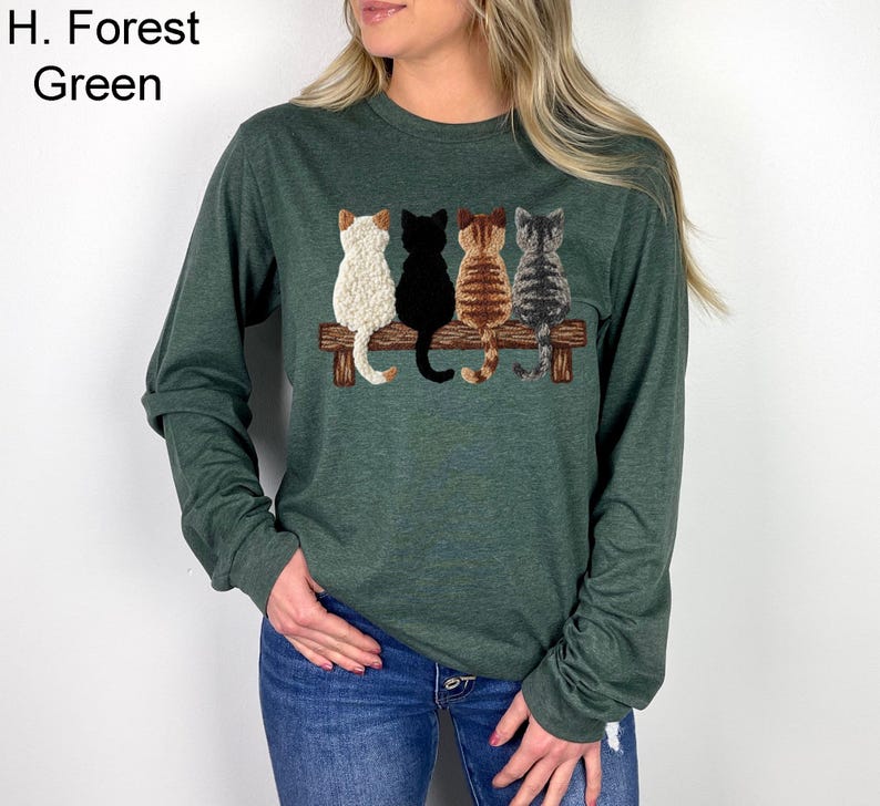 May include: Forest green long-sleeved top with an embroidered design of four cats on a wooden bench. The cats are white, black, brown, and grey. The shirt has a crew neck and is made of a soft material.