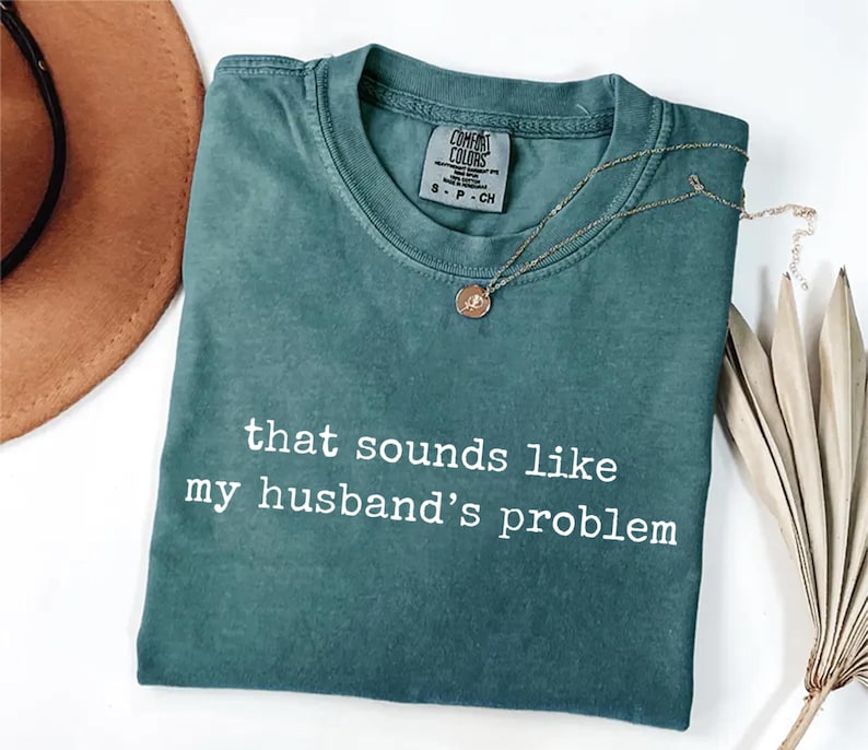 May include: A teal Comfort Colors t-shirt with the phrase "that sounds like my husband's problem" in white lettering. A gold necklace with a pendant rests on the shirt. A brown hat and dried plant are in the background.