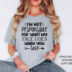 K&ouml;nnte beinhalten: Ein hellgraues T-Shirt mit schwarzem Text, der "Ich bin nicht verantwortlich f&uuml;r das, was mein Gesicht tut, wenn du sprichst" lautet, mit einem schwarzen Pfeil, der nach rechts zeigt. Das Shirt ist aus einem weichen, bequemen Material.