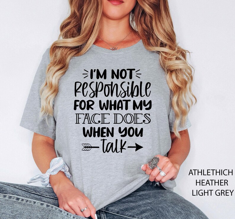 K&ouml;nnte beinhalten: Ein hellgraues T-Shirt mit schwarzem Text, der "Ich bin nicht verantwortlich f&uuml;r das, was mein Gesicht tut, wenn du sprichst" lautet, mit einem schwarzen Pfeil, der nach rechts zeigt. Das Shirt ist aus einem weichen, bequemen Material.