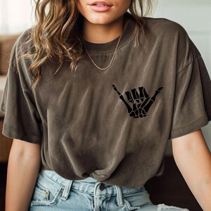 Pode incluir: Uma camiseta marrom com uma m&atilde;o de esqueleto preta fazendo um sinal shaka. A camiseta &eacute; feita de um material macio e confort&aacute;vel.
