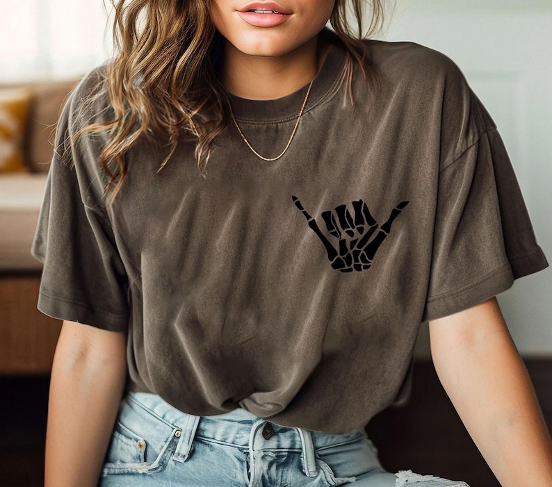 Pode incluir: Uma camiseta marrom com uma m&atilde;o de esqueleto preta fazendo um sinal shaka. A camiseta &eacute; feita de um material macio e confort&aacute;vel.