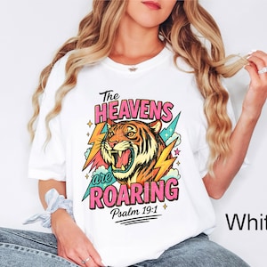 Puede incluir: Camiseta blanca con un gr&aacute;fico colorido de un tigre rugiendo, rayos y el texto "The Heavens are Roaring" y "Psalm 19:1". El dise&ntilde;o presenta colores rosa, naranja, amarillo y turquesa.