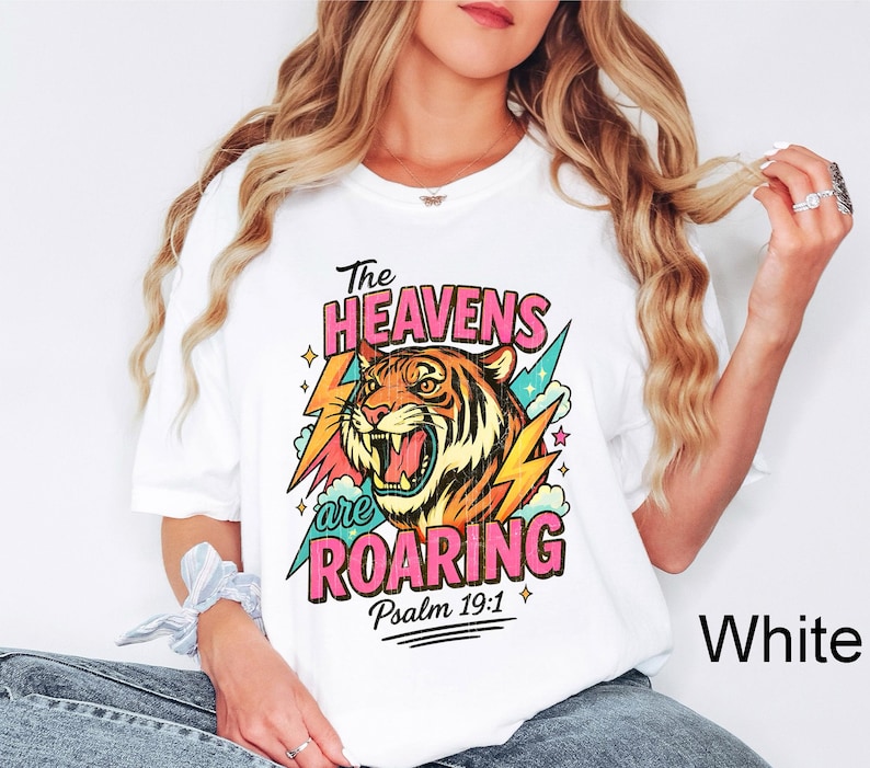 Puede incluir: Camiseta blanca con un gr&aacute;fico colorido de un tigre rugiendo, rayos y el texto "The Heavens are Roaring" y "Psalm 19:1". El dise&ntilde;o presenta colores rosa, naranja, amarillo y turquesa.