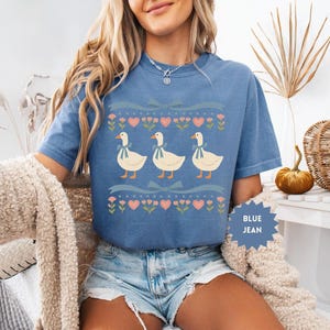 Pode incluir: Camiseta azul jeans com um design l&uacute;dico de tr&ecirc;s gansos com la&ccedil;os, em um fundo de cora&ccedil;&otilde;es, flores e fitas. A camiseta tem mangas curtas e um corte folgado.