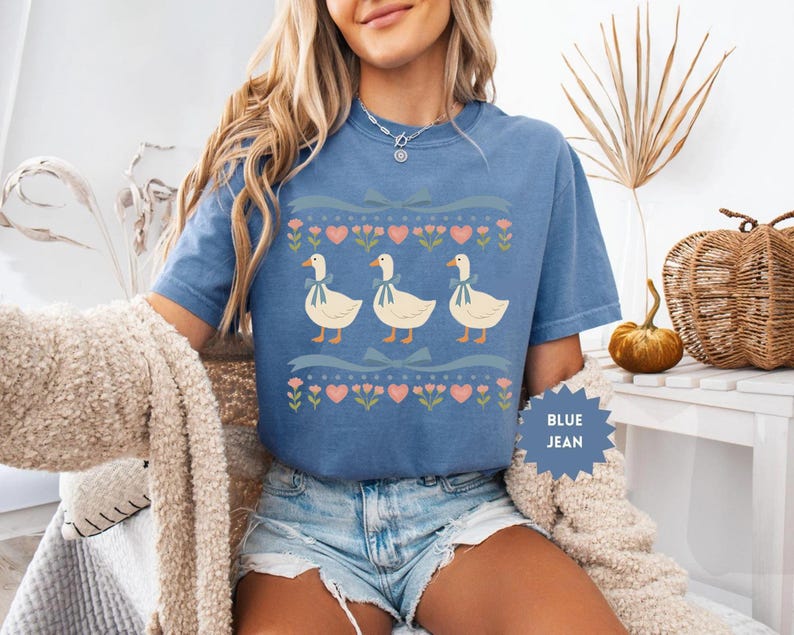 Pode incluir: Camiseta azul jeans com um design l&uacute;dico de tr&ecirc;s gansos com la&ccedil;os, em um fundo de cora&ccedil;&otilde;es, flores e fitas. A camiseta tem mangas curtas e um corte folgado.