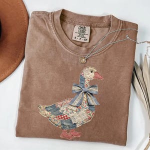 Peut inclure: Un t-shirt marron Comfort Colors avec un motif d'oie appliqu&eacute;. L'oie a un corps en patchwork, un n&oelig;ud bleu et un bec et des pieds &agrave; carreaux rouges et blancs. Un collier en argent avec un petit pendentif est drap&eacute; sur le t-shirt.