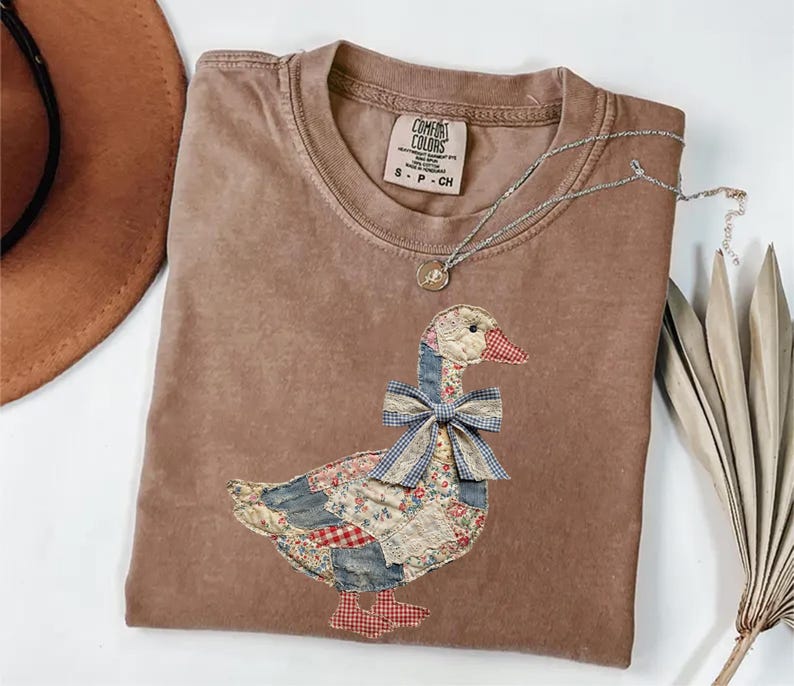 Peut inclure: Un t-shirt marron Comfort Colors avec un motif d'oie appliqu&eacute;. L'oie a un corps en patchwork, un n&oelig;ud bleu et un bec et des pieds &agrave; carreaux rouges et blancs. Un collier en argent avec un petit pendentif est drap&eacute; sur le t-shirt.
