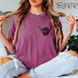 Pode incluir: Uma mulher vestindo uma camiseta roxa com uma m&atilde;o de esqueleto preta fazendo um sinal shaka. A camiseta tem a palavra "BERRY" impressa no canto superior direito.