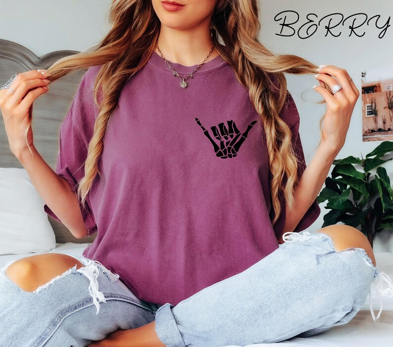 Pode incluir: Uma mulher vestindo uma camiseta roxa com uma m&atilde;o de esqueleto preta fazendo um sinal shaka. A camiseta tem a palavra "BERRY" impressa no canto superior direito.