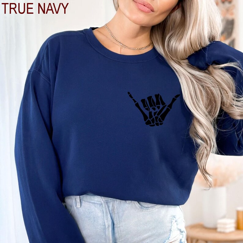 Pode incluir: Uma camisa de moletom azul marinho com uma m&atilde;o de esqueleto preta fazendo um sinal shaka. O texto "TRUE NAVY" &eacute; vis&iacute;vel no canto superior esquerdo da imagem.