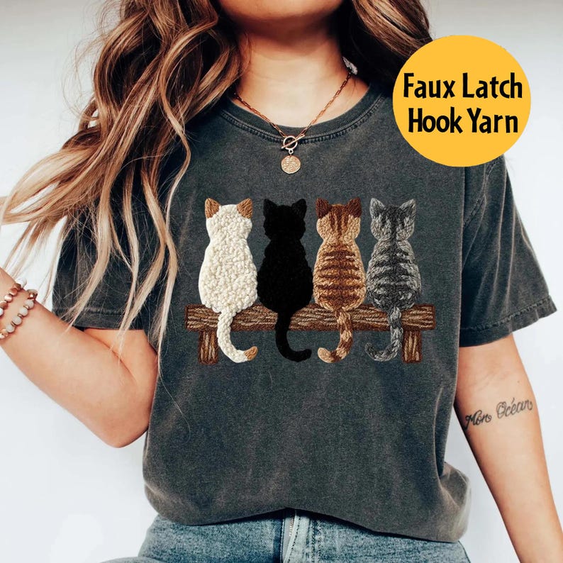 May include: Dark grey t-shirt with an embroidered design of four cats on a wooden bench. The cats are white, black, brown, and grey. The text "Faux Latch Hook Yarn" is in a yellow circle.