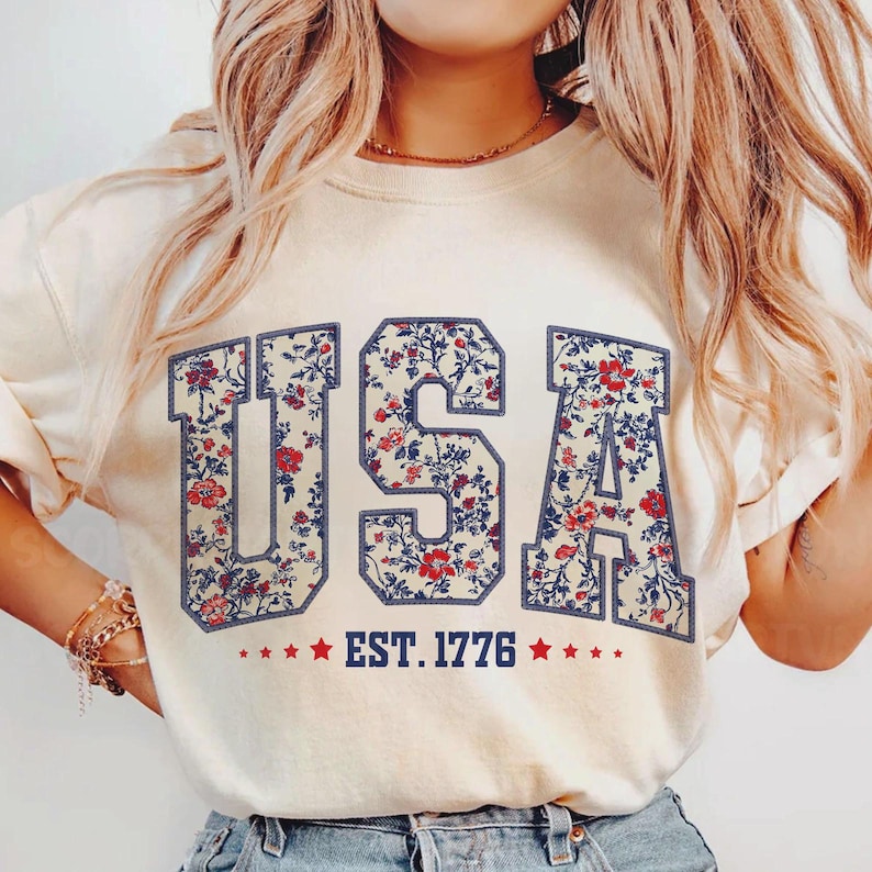 May include: Cream-colored t-shirt with the word "USA" in a floral pattern. The letters are outlined in navy blue, with the text "EST. 1776" and red stars below. A patriotic design.
