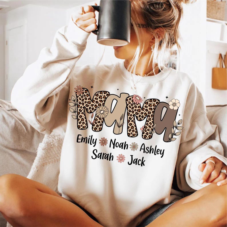 May include: Cream-colored sweatshirt with the word "Mama" in a leopard print and brown font. The names Emily, Noah, Sarah, Ashley, and Jack are printed below the word "Mama" with floral accents.