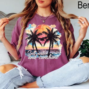 Op de afbeelding: Een paars T-shirt met een afbeelding van palmbomen en een zonsondergang. De tekst "Salt water and sunshine" staat op het shirt gedrukt.