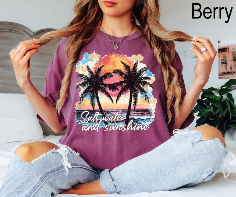 Op de afbeelding: Een paars T-shirt met een afbeelding van palmbomen en een zonsondergang. De tekst "Salt water and sunshine" staat op het shirt gedrukt.