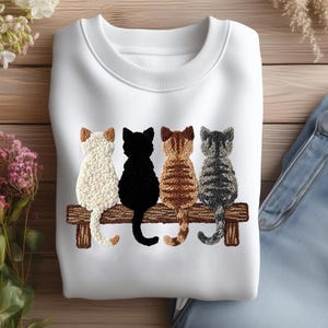 May include: White sweatshirt with an embroidered design of four cats on a wooden bench. The cats are cream, black, brown, and grey. The sweatshirt is made of a soft material, perfect for casual wear.