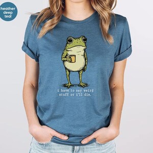 May include: Heather deep teal t-shirt featuring a cartoon frog holding a mug. The frog is green and beige, with the text "I have to say weird stuff or I'll die." printed below the frog. The shirt is a solid color.