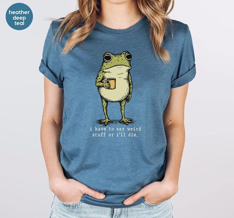May include: Heather deep teal t-shirt featuring a cartoon frog holding a mug. The frog is green and beige, with the text "I have to say weird stuff or I'll die." printed below the frog. The shirt is a solid color.