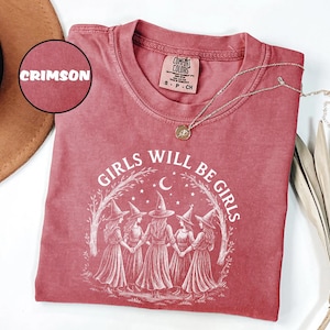 May include: Crimson-coloured t-shirt with the text "GIRLS WILL BE GIRLS" and an illustration of five witches in a circle. The shirt has a crew neck and short sleeves. A gold necklace with a pendant is on the shirt.