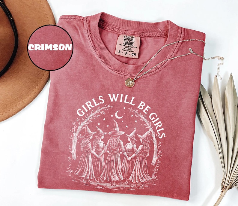 May include: Crimson-coloured t-shirt with the text "GIRLS WILL BE GIRLS" and an illustration of five witches in a circle. The shirt has a crew neck and short sleeves. A gold necklace with a pendant is on the shirt.