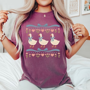 Pode incluir: Uma camiseta roxa com um design l&uacute;dico de tr&ecirc;s gansos brancos com la&ccedil;os azuis, em um fundo de cora&ccedil;&otilde;es, flores e fitas azuis. O design &eacute; centralizado na camiseta, criando uma est&eacute;tica charmosa e divertida.