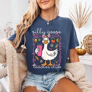 Puede incluir: Camiseta azul marino con un dise&ntilde;o de ganso de dibujos animados. El ganso sostiene una bebida rosa y lleva un lazo. El texto de la camiseta dice "silly goose teacher club".