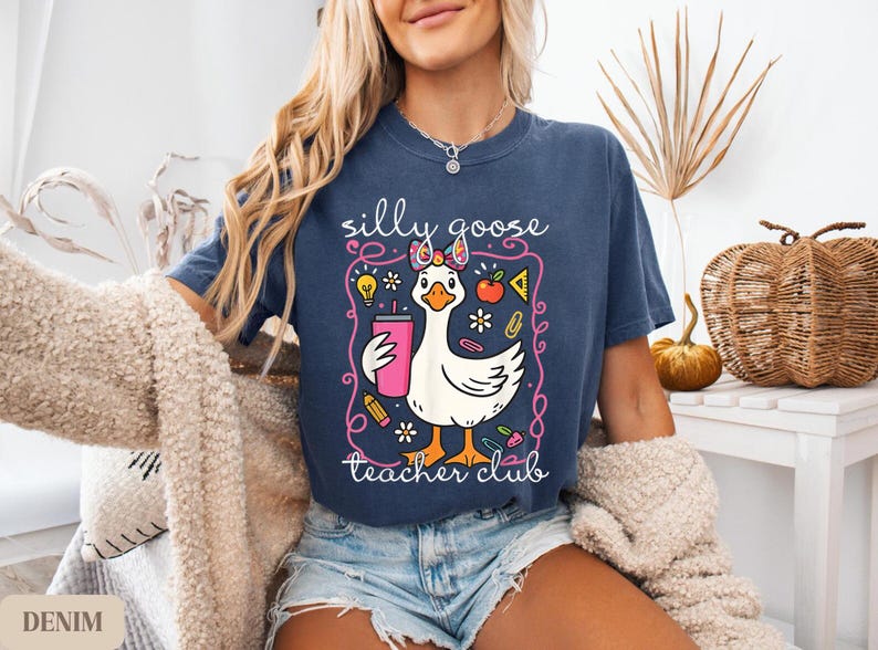Puede incluir: Camiseta azul marino con un dise&ntilde;o de ganso de dibujos animados. El ganso sostiene una bebida rosa y lleva un lazo. El texto de la camiseta dice "silly goose teacher club".