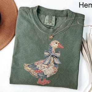 Peut inclure: Un t-shirt vert Comfort Colors avec un motif d'oie en patchwork. L'oie a un bec rouge, un n&oelig;ud bleu et un corps fait de divers motifs de tissu. Un collier en argent avec un pendentif repose sur le t-shirt. Le mot "Hemp" est dans le coin sup&eacute;rieur droit.