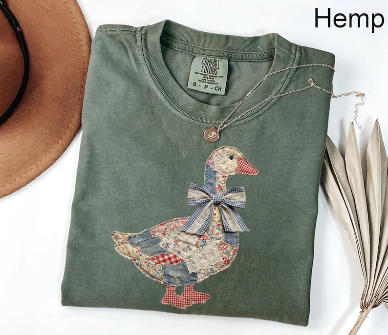 Peut inclure: Un t-shirt vert Comfort Colors avec un motif d'oie en patchwork. L'oie a un bec rouge, un n&oelig;ud bleu et un corps fait de divers motifs de tissu. Un collier en argent avec un pendentif repose sur le t-shirt. Le mot "Hemp" est dans le coin sup&eacute;rieur droit.