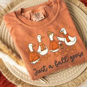 Puede incluir: Camiseta naranja Comfort Colors con una ilustraci&oacute;n de cuatro gansos vestidos con atuendos oto&ntilde;ales. Los gansos llevan bufandas, su&eacute;teres y un sombrero, con el texto "Just a fall goose" debajo. La camiseta es de material suave.