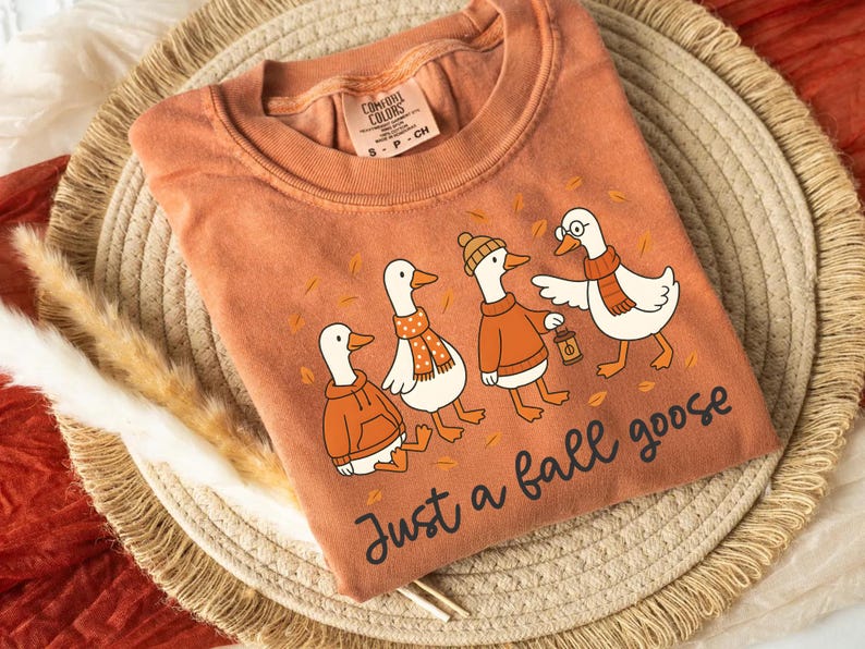 Puede incluir: Camiseta naranja Comfort Colors con una ilustraci&oacute;n de cuatro gansos vestidos con atuendos oto&ntilde;ales. Los gansos llevan bufandas, su&eacute;teres y un sombrero, con el texto "Just a fall goose" debajo. La camiseta es de material suave.