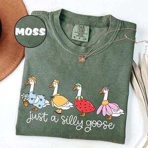 Puede incluir: Camiseta verde musgo con un dise&ntilde;o caprichoso de cuatro gansos vestidos con varios atuendos, incluyendo un vestido floral y un disfraz de fresa. La frase "just a silly goose" est&aacute; escrita en cursiva debajo de los gansos. Un collar dorado est&aacute; sobre la camiseta.