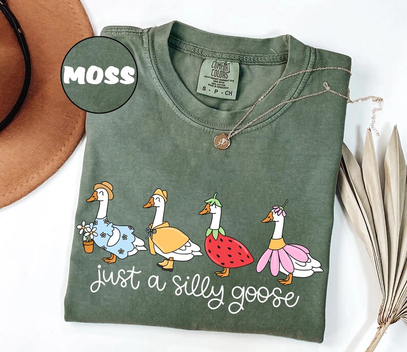 Puede incluir: Camiseta verde musgo con un dise&ntilde;o caprichoso de cuatro gansos vestidos con varios atuendos, incluyendo un vestido floral y un disfraz de fresa. La frase "just a silly goose" est&aacute; escrita en cursiva debajo de los gansos. Un collar dorado est&aacute; sobre la camiseta.