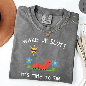 Puede incluir: Una camiseta gris Comfort Colors con el texto "WAKE UP SLUTS IT'S TIME TO SIN". El dise&ntilde;o incluye un sol sonriente con gafas de sol, un gusano rojo y flores azules. Un collar plateado con un colgante est&aacute; sobre la camiseta.