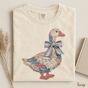 Peut inclure: T-shirt ivoire avec un motif d'oie en patchwork. L'oie a un corps en denim bleu, des accents floraux et vichy rouge, et un n&oelig;ud vichy bleu. Le t-shirt est en mati&egrave;re douce et a un col rond.