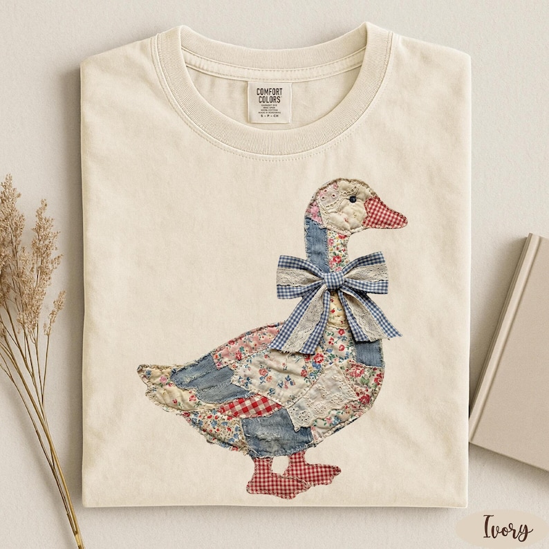 Peut inclure: T-shirt ivoire avec un motif d'oie en patchwork. L'oie a un corps en denim bleu, des accents floraux et vichy rouge, et un n&oelig;ud vichy bleu. Le t-shirt est en mati&egrave;re douce et a un col rond.