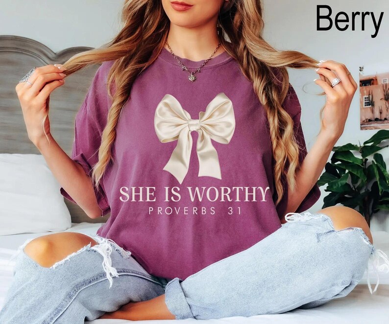 May include: A mauve t-shirt with a large white bow and the text "She is Worthy Proverbs 31" printed on the front.