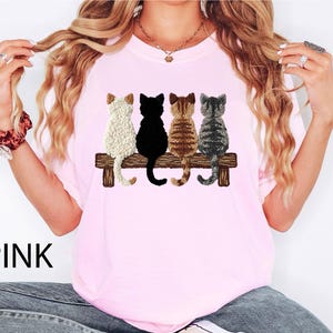 May include: Pale pink t-shirt with an embroidered design of four cats on a wooden bench. The cats are white, black, brown, and grey. The word "PINK" is printed in black at the bottom left of the shirt.