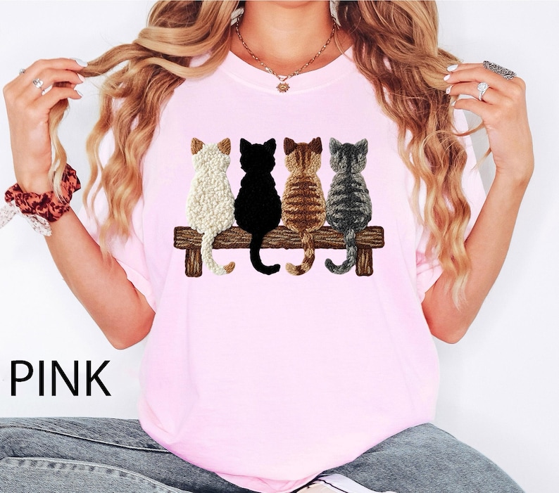 May include: Pale pink t-shirt with an embroidered design of four cats on a wooden bench. The cats are white, black, brown, and grey. The word "PINK" is printed in black at the bottom left of the shirt.