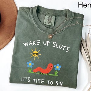 Puede incluir: Camiseta verde oliva con el texto "WAKE UP SLUTS IT'S TIME TO SIN". El dise&ntilde;o incluye un sol sonriente con gafas de sol, un gusano rojo y flores azules. Un collar plateado con un colgante est&aacute; sobre la camiseta.