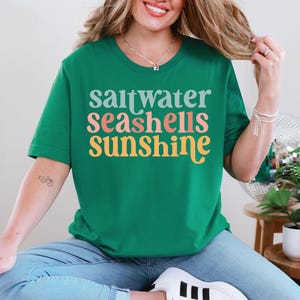 May include: A green t-shirt featuring the words "saltwater seashells sunshine" in a retro font. The text is in teal, coral, and yellow. The t-shirt has a crew neck and is a solid green colour.