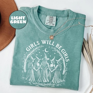 May include: Light green t-shirt featuring the text "GIRLS WILL BE GIRLS" and an illustration of five witches holding hands. The shirt has a crew neck and short sleeves. A silver necklace with a pendant is draped across the shirt.