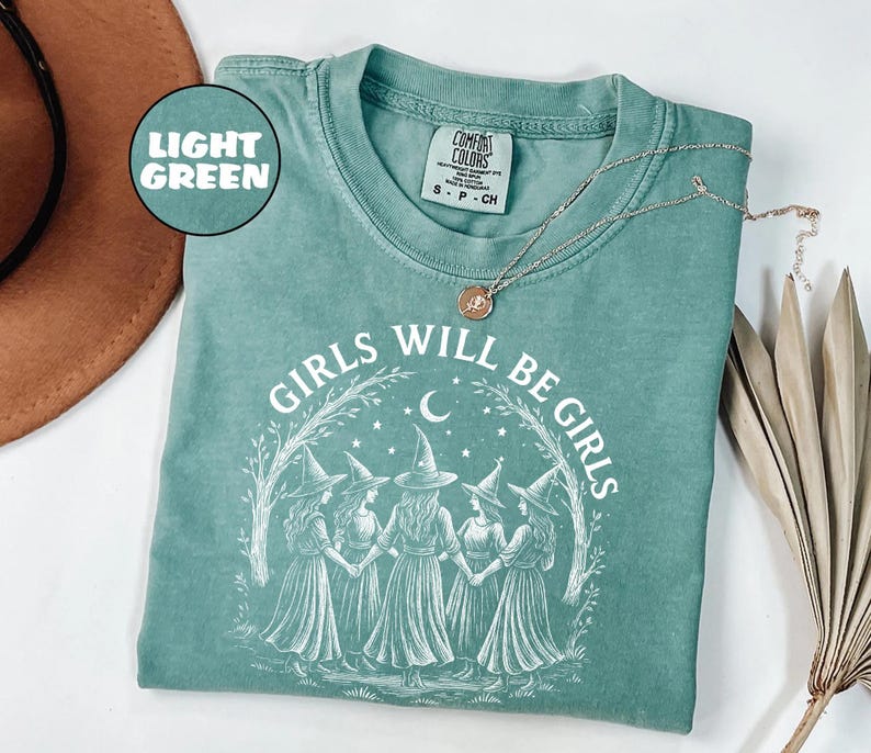 May include: Light green t-shirt featuring the text "GIRLS WILL BE GIRLS" and an illustration of five witches holding hands. The shirt has a crew neck and short sleeves. A silver necklace with a pendant is draped across the shirt.