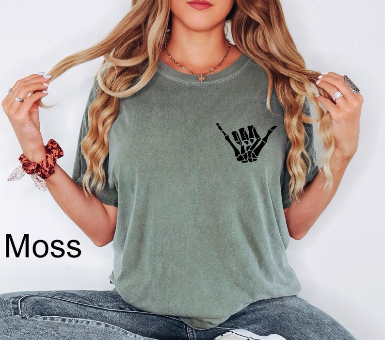 Pode incluir: Uma mulher vestindo uma camiseta verde musgo com um gr&aacute;fico de m&atilde;o de esqueleto preto "hang loose". A camiseta tem um caimento relaxado e mangas curtas.